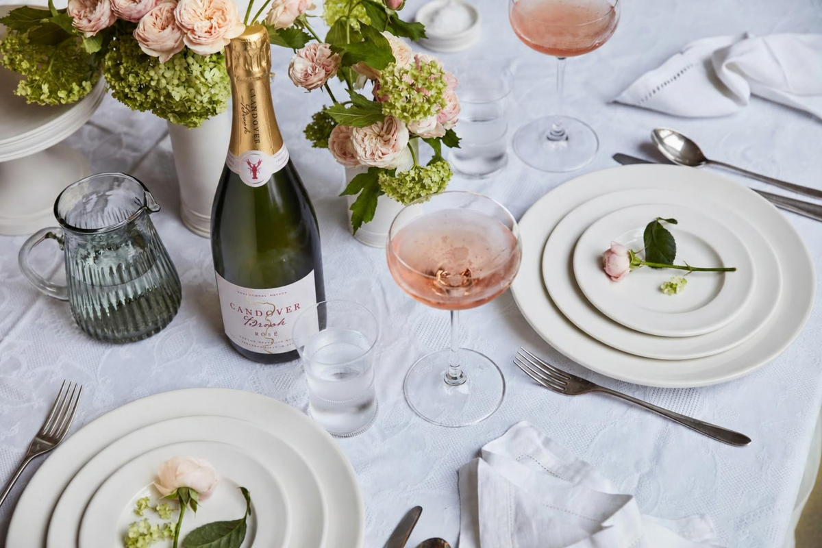 A styled table setting with white plates, silver cutlery, a bottle of Candover Brook Rosé sparkling wine, pink rose wine in stemmed glasses, and floral arrangements on a white tablecloth.