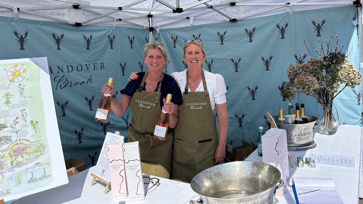 Two women smiling behind a table under a white canopy tent, holding bottles of sparkling wine. The backdrop features a teal banner with the brand name 'Candover Brook' and black crayfish logos.