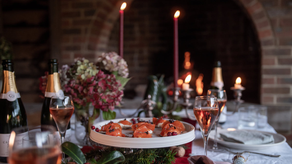 A festive dining table set in front of a brick fireplace, featuring champagne bottles, filled wine glasses, candles, and a white cake stand holding pastries. The foreground includes greenery and red berries, creating a warm, celebratory atmosphere.