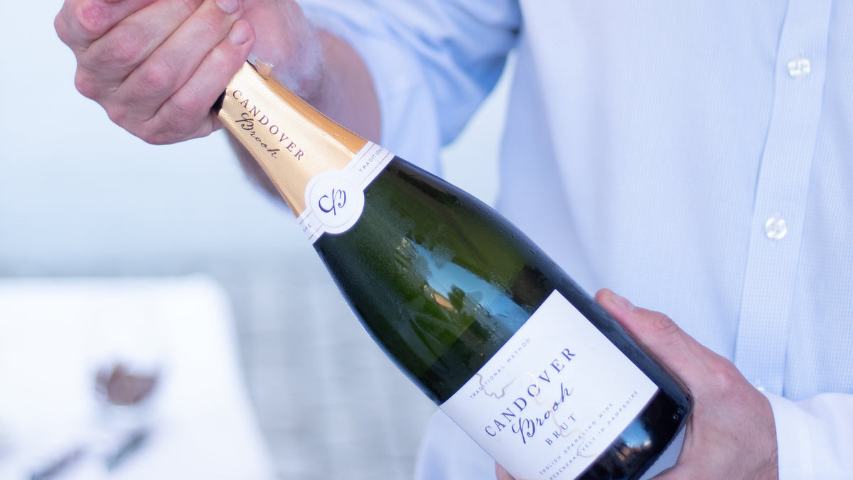 A person in a light blue dress shirt holding and opening a bottle of Candover Brook Brut english sparkling wine.