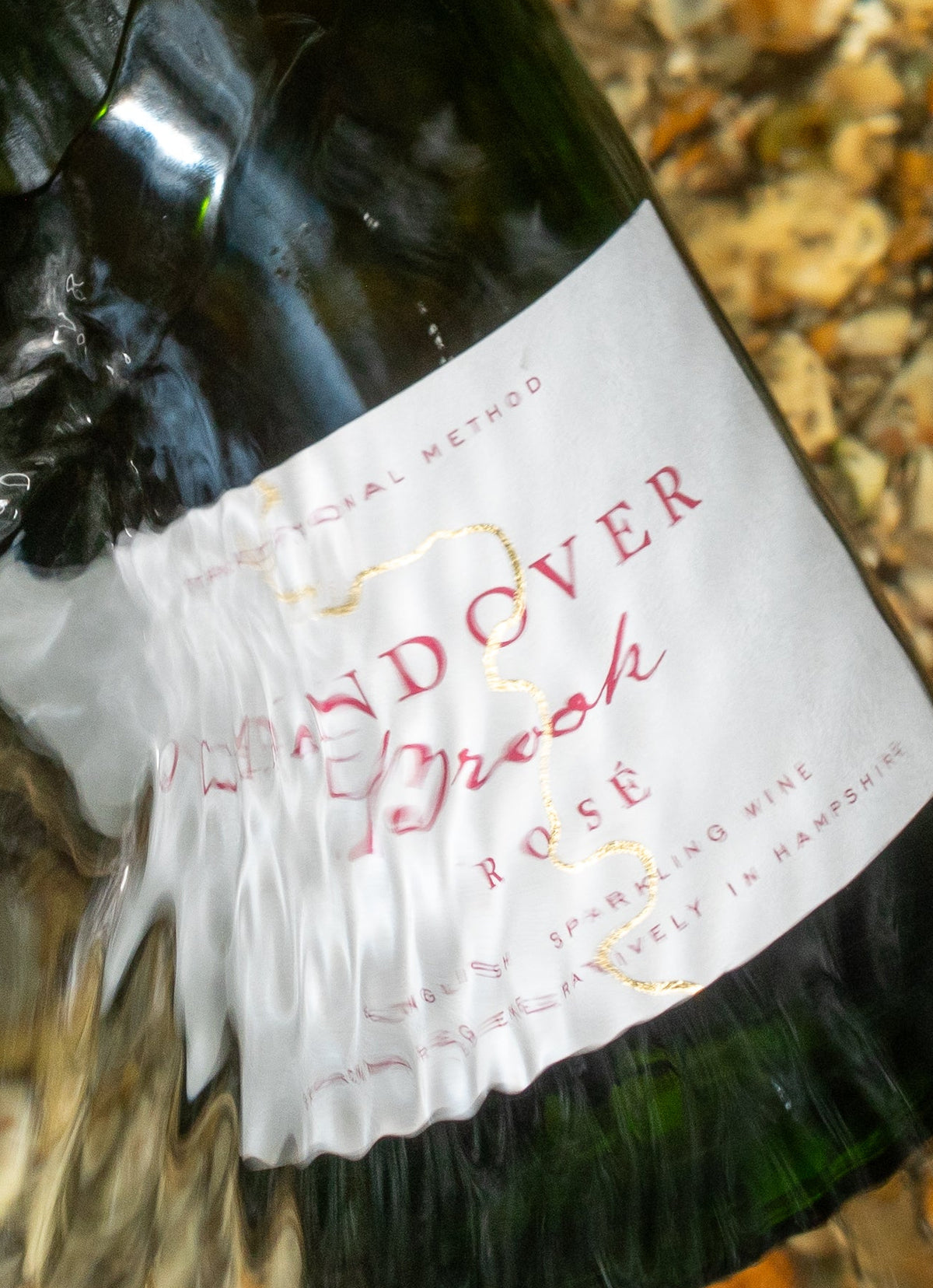 A bottle of Candover Brook Sparkling Rosé Wine resting on wet river stones and water, with clear water ripples flowing around the dark green glass and gold foil.