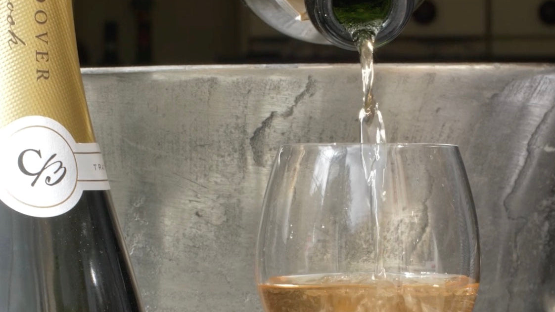 A bottle of sparkling wine or champagne with a gold foil label is partially visible on the left, while another bottle pours pale golden liquid into a clear wine glass against a metallic gray background.