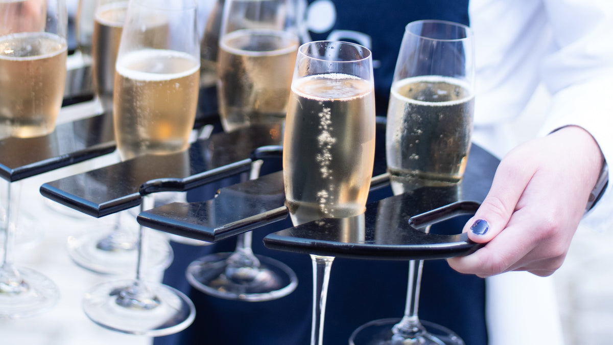 A close-up of a person in a white shirt and navy apron holding a black tray with several champagne flutes filled with candover brook brut nv sparkling wine 